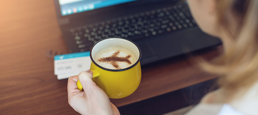 Mujer con una taza de café con espuma y una silueta de un avión usando el ordenador con unos pasajes encima.